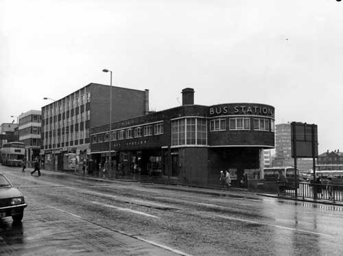 Black and white photograph showing an urban street, with a bus station in the centre and a row of office blocks to the left of centre