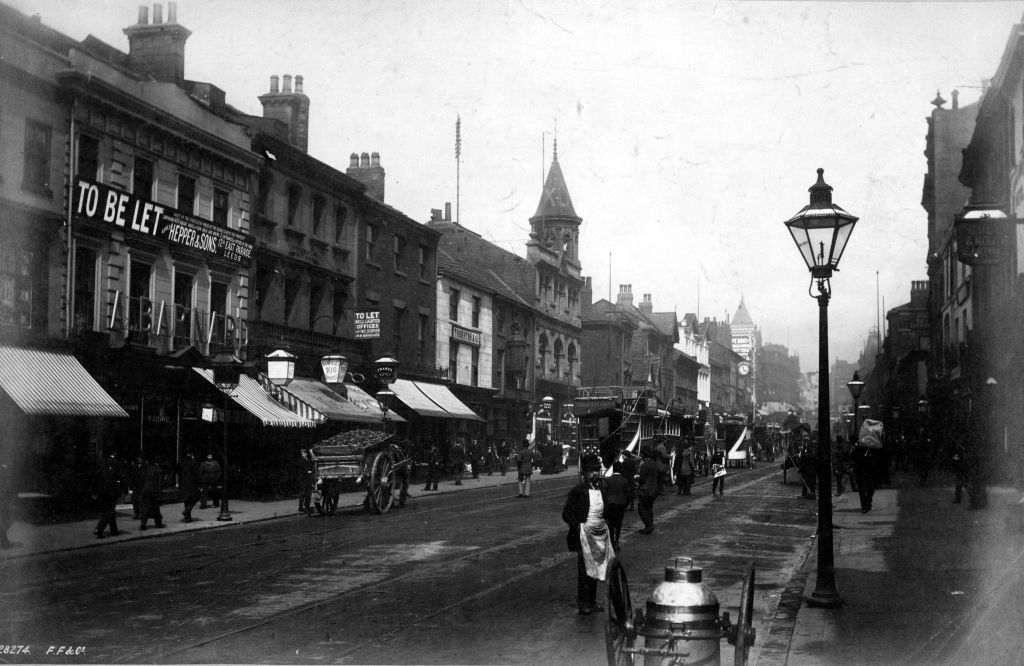 A black and white photograph showing Briggate. People are walking on pavements and trams and horse and carts can be seen on the road. 