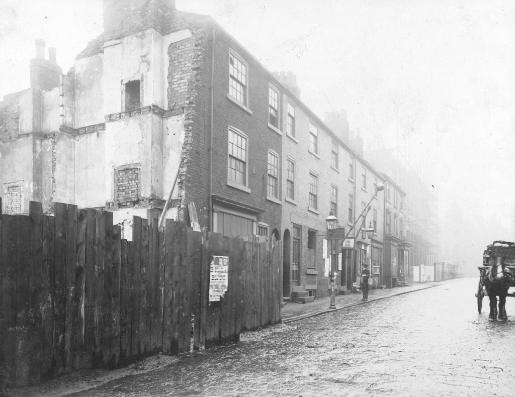 Black and white view of street with a horse and cart in view. A building has been demolished and fencing has been placed in front of it.