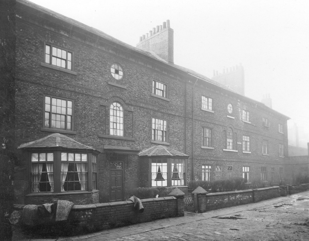 Black and white photograph showing a row of three story houses with bay windows and a small wall running outside each. A couple of rolls of carpet are laying over the walls.