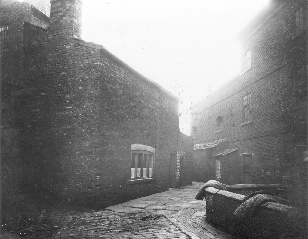 Black and white image showing buildings perpendicular to each other. A short wall in the foreground has some rolls of carpet laying over it.