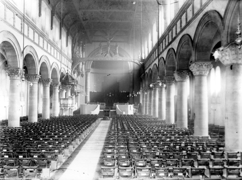Black and white view of the inside of a church. Rows of pews are visible and the alter is at the far end of the image.