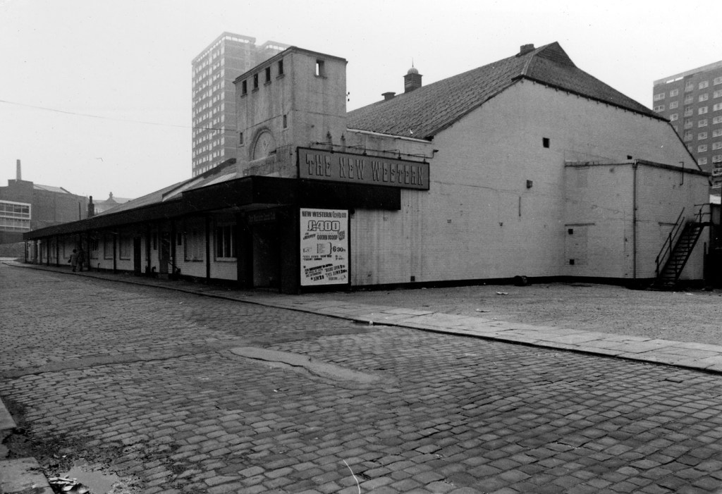 Black and white photo showing an old cinema building. The road outside it is cobbled. A large block of flats is in the distance.