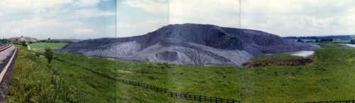 Colour photograph showing green grass and a hill of a mine behind it.
