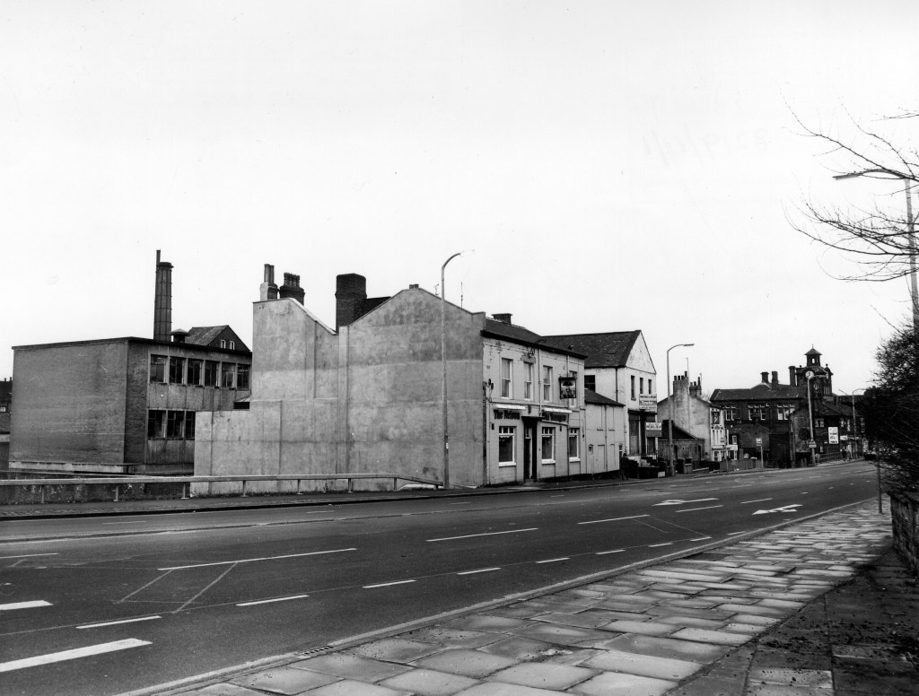 Black and white photo with road in the foreground and some buildings to the left of the road. One of these is The Nelson public house.
