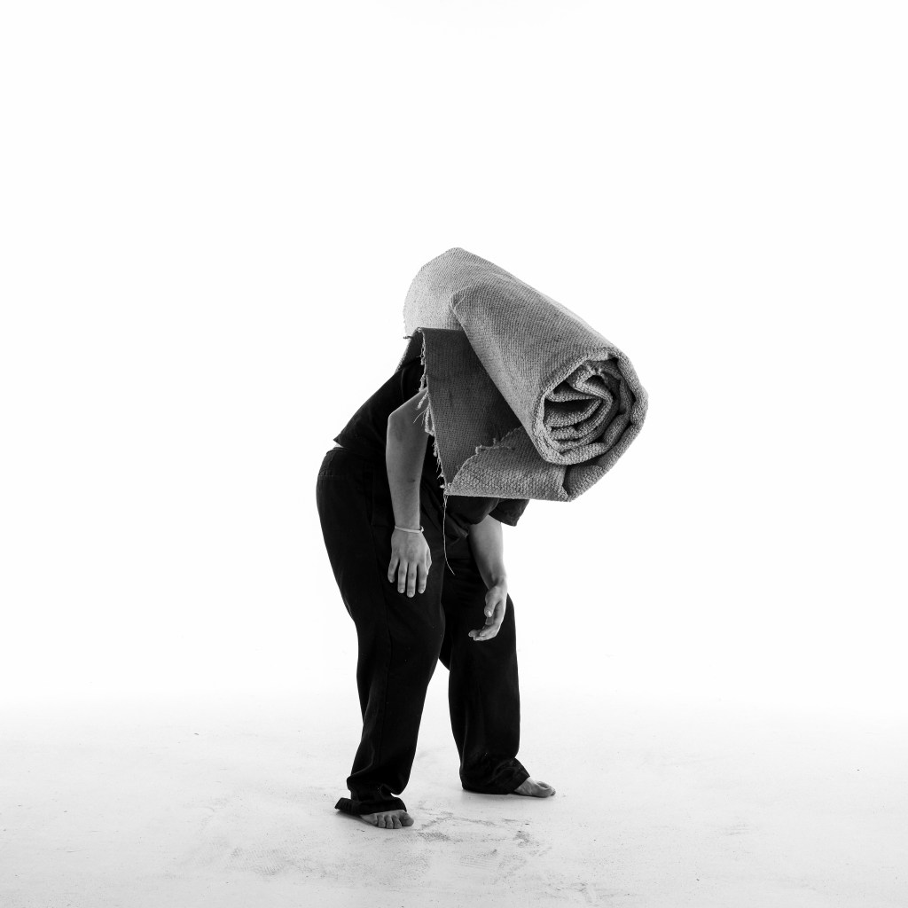 Black and white photograph of a barefoot man with dark clothing with a roll of carpet over his shoulder. His face is obscured by the carpet.