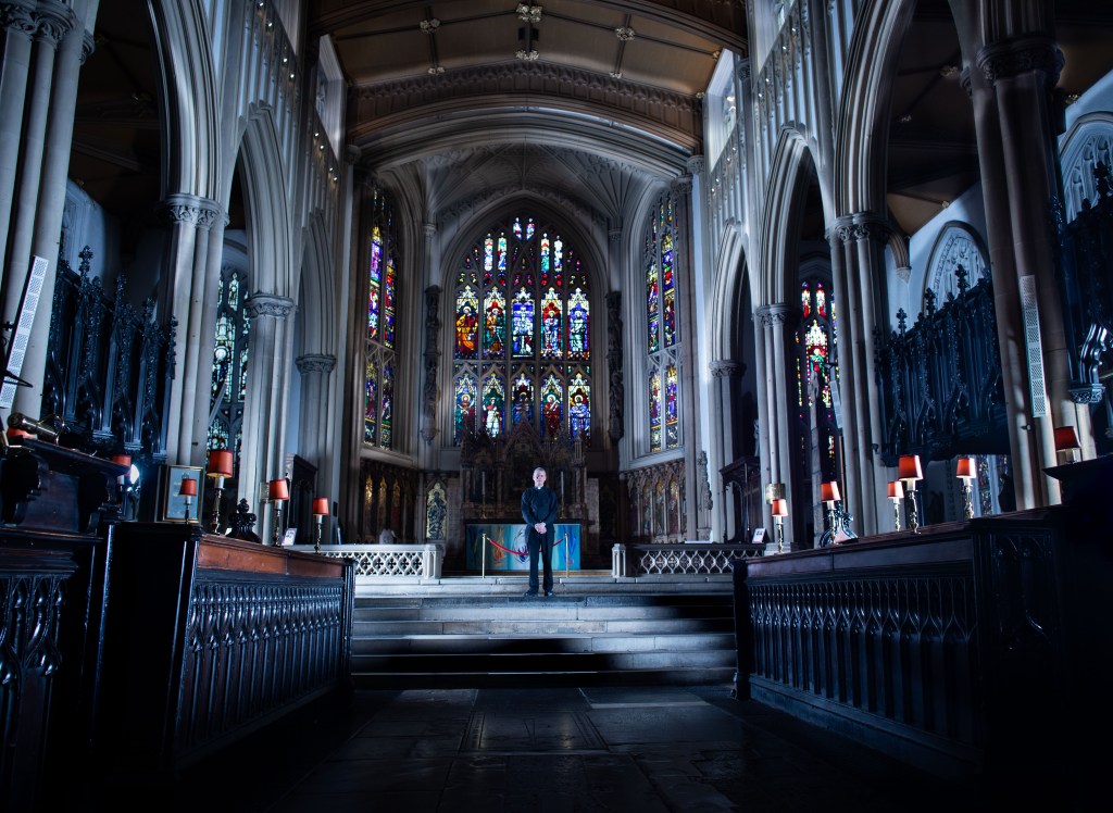 Colour photograph of the interior of a church. The alter is in view with lots of stain glass windows surrounding it. A vicar stands in front of the alter.