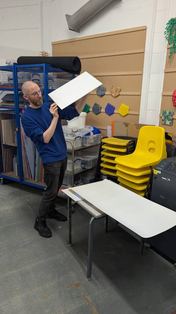 A man holding up a piece of rectangular card. In front of him is a table with a large piece of paper laid across it.