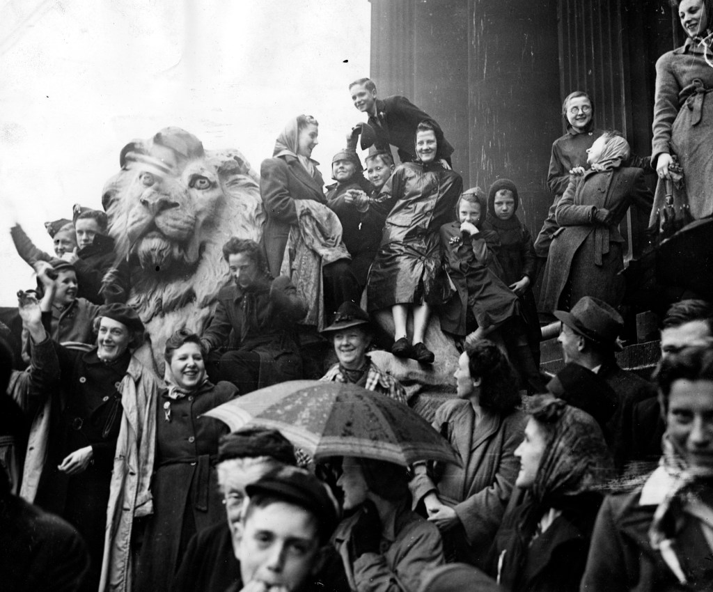 A group of people, mostly women, stand on the steps of Leeds Town Hall to celebrate the ending of the war in Europe. A stone lion can be seen amongst the crowd.